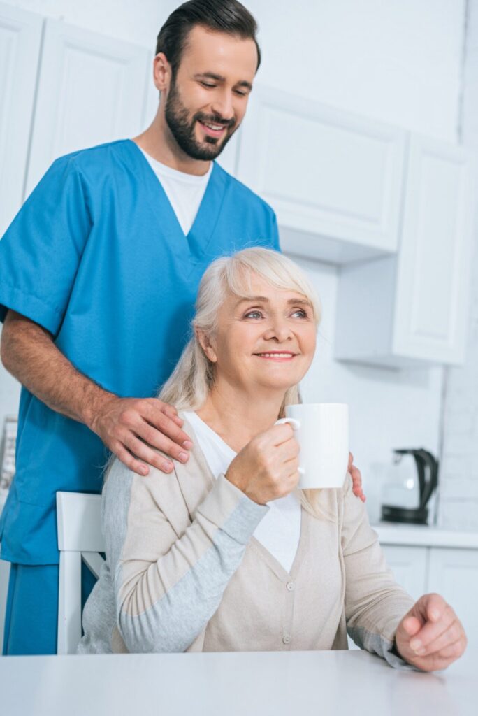 smiling male caregiver looking at senior woman drinking tea in kitchen